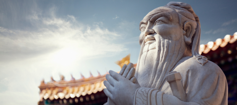 Stone statue of Confucius, hands clasped, against a temple backdrop.