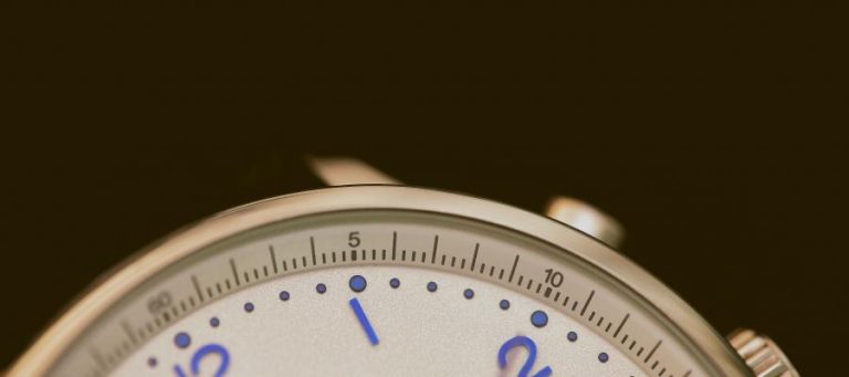 Close-up of a watch face; silver casing, white face, blue markers.