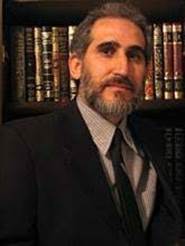 Portrait of a man with a beard, wearing a suit and tie, in front of a bookshelf filled with books.
