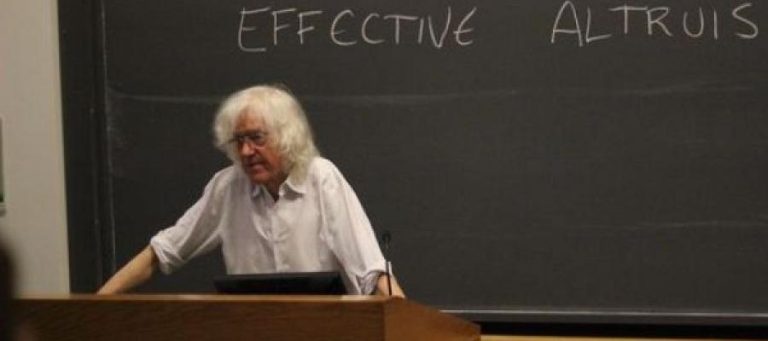 Older man with white hair lecturing at a blackboard with "Effective Altruism" written on it.