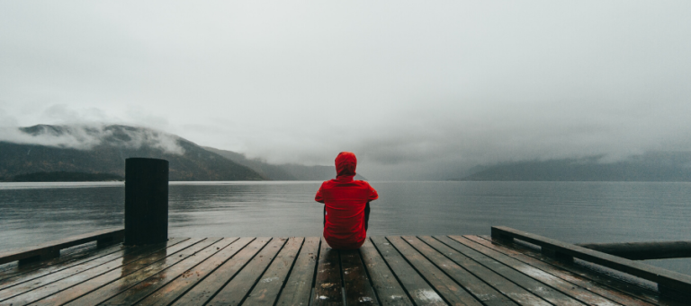 Person in red hooded jacket sits on a dock overlooking a calm lake and mountains.