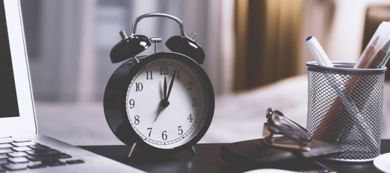 Black alarm clock showing almost noon, next to a laptop and glasses.