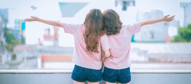 Two women stand back-to-back, arms outstretched, gazing at a cityscape.