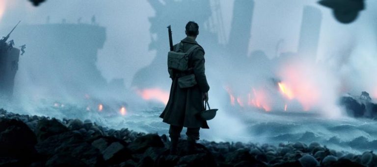 A lone soldier stands on a desolate beach, overlooking burning ships at sea.