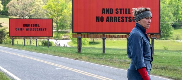 Woman stands by roadside billboards reading "How come, Chief Willoughby?" and "And still no arrests?".