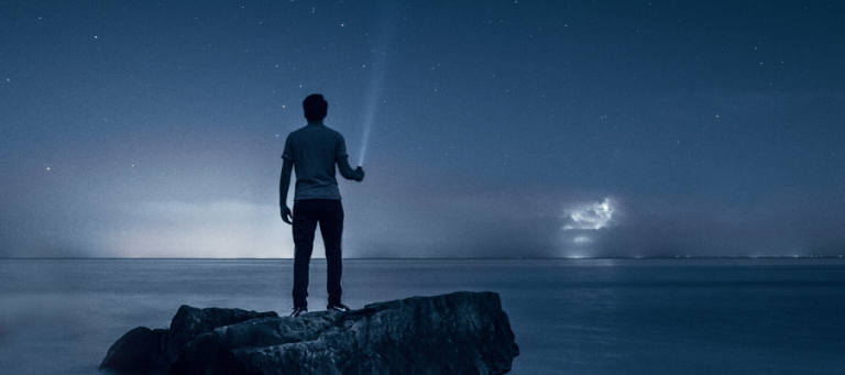 Silhouetted person on a rock by the sea at night, holding a flashlight, gazing at the stars.