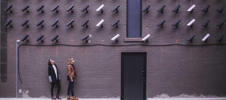 Two women stand before a wall of surveillance cameras, contemplating privacy in the digital age.