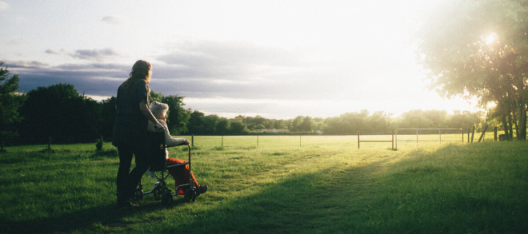 Woman pushing elderly person in wheelchair across grassy field.