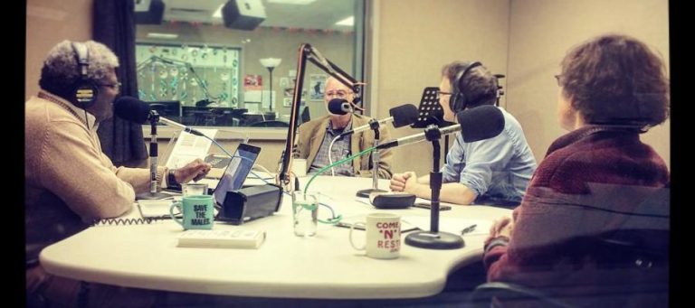 Four people participate in a radio show recording, seated around a table with microphones.