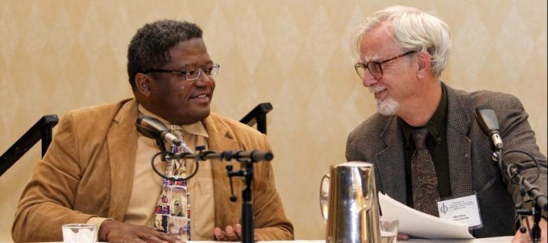 Two men in suits converse at a conference, microphones present.