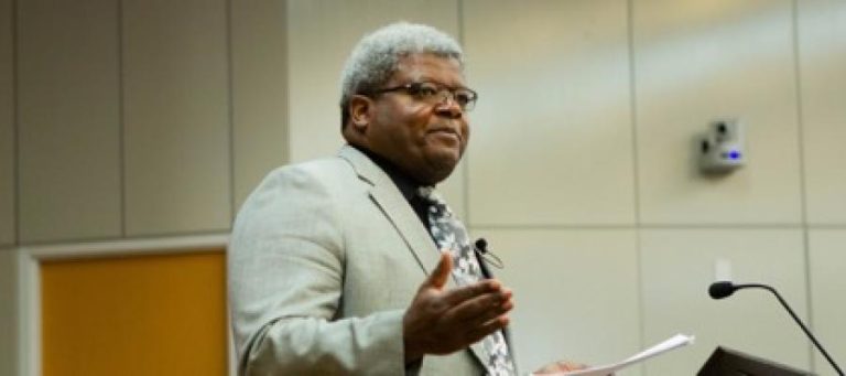Man in suit speaking at podium, expressing jovial emotion.