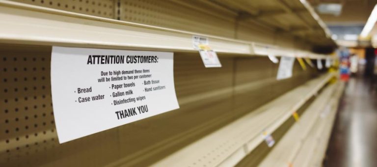Empty grocery store shelves with a sign limiting purchases due to high demand. The sign lists bread, water, milk, and sanitizing products.