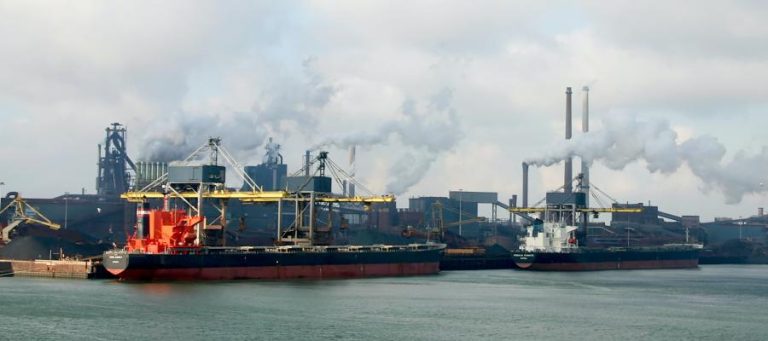 Cargo ships at industrial port with smokestacks emitting pollution.