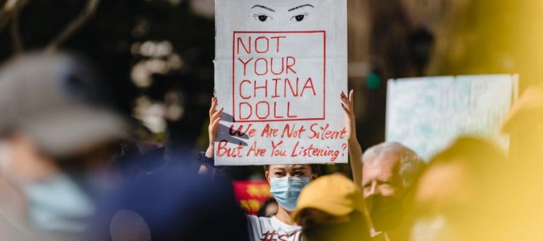 Protestor holds sign reading "Not Your China Doll. We Are Not Silent. But Are You Listening?