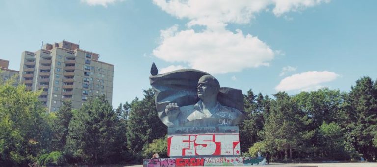 Graffiti-covered bust of a man, possibly a leader, with a raised fist. A residential building is visible in the background.