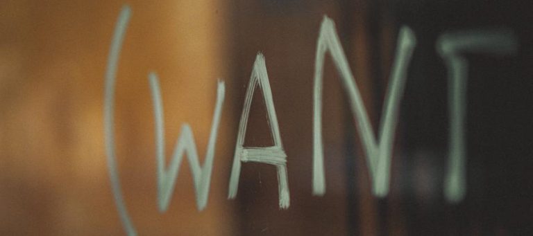 Handwritten word "WANT" on a dark surface, reflecting a blurred background.
