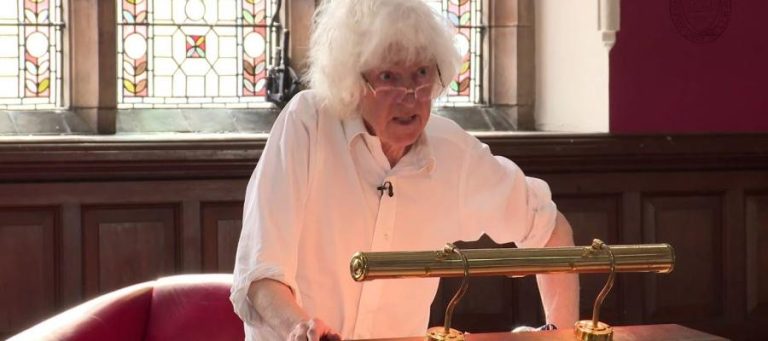 Elderly woman with white hair, wearing a white shirt, speaks at a lectern in a wood-paneled room.