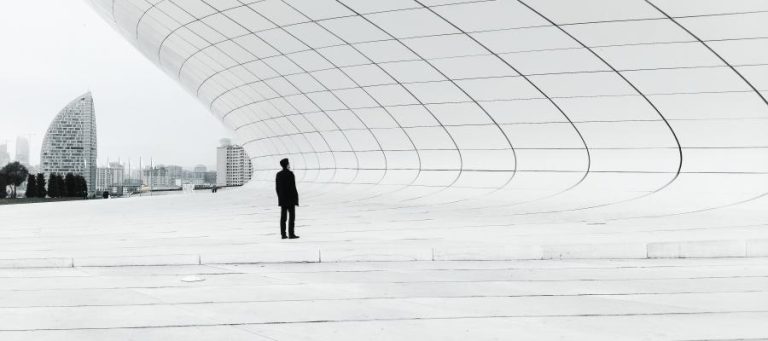 Man in silhouette stands before a modern, curved building, city skyline in background.