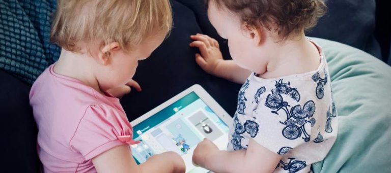Two toddlers share a tablet, engrossed in a children's app.