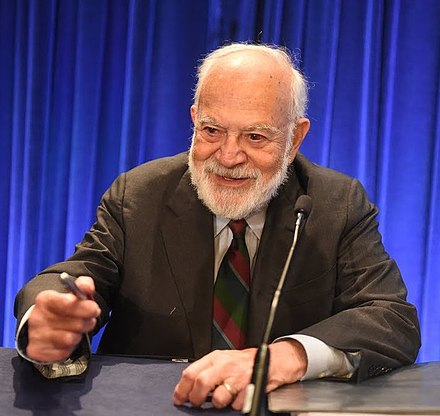 Elderly man with beard, speaking at a podium, holding a pen. Truth, BS.