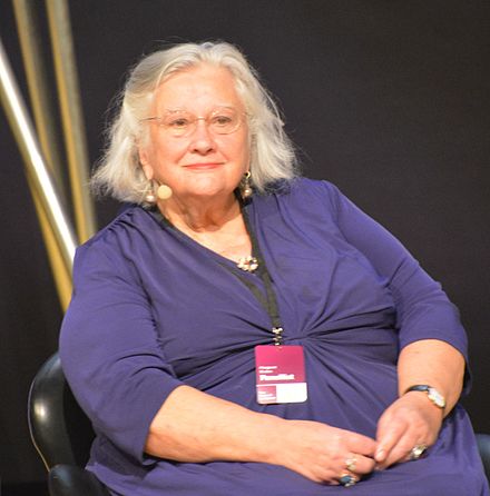 Older woman with short gray hair, wearing a purple dress, sits and smiles. Creativity.