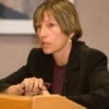 A woman with short brown hair sits at a podium, appearing serious.