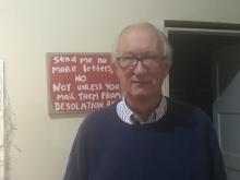 Older man wearing glasses, standing in front of a sign that says "Send me no more letters, no, not unless you mail them from Desolation Row.