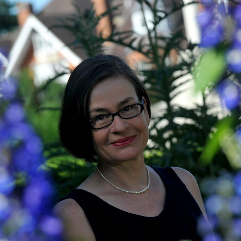 Portrait of a woman with short dark hair and glasses, wearing a black dress and pearl necklace.