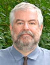 Headshot of a man with graying hair and beard, wearing a light blue shirt and tie.