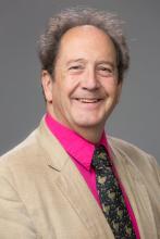 Headshot of a smiling man in a beige jacket and pink shirt.