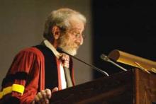 Elderly man in academic regalia giving a speech at a podium.