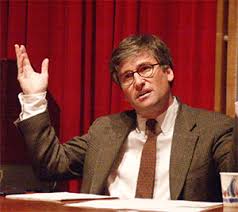 Man in suit and glasses gesturing during a presentation.