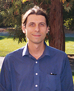 Headshot of a man with shoulder-length brown hair, wearing a blue collared shirt.