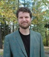 Headshot of a man with short brown hair and a beard, wearing a green jacket.
