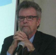 Man with glasses speaking at a microphone, discussing finding meaning in a material world.