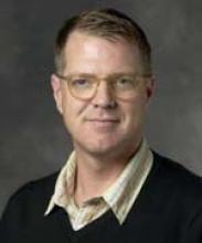 Headshot of a middle-aged man with glasses, wearing a dark v-neck sweater over a collared shirt.