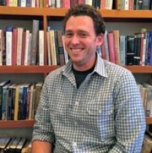 Smiling man in a plaid shirt, sitting in front of bookshelves.