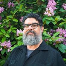 Portrait of a man with a gray beard, wearing glasses, in front of rhododendrons.