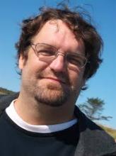 Headshot of a man with glasses and a goatee, smiling outdoors.